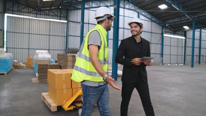 Male procurement officer in black business suit has friendly conversation with warehouse worker.