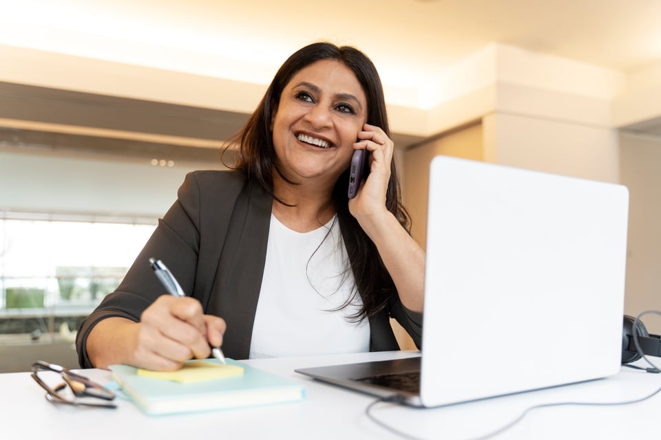 Procurement manager sits at desk with her laptop computer, talking on smartphone to GPO about ways to reduce telecom expenses