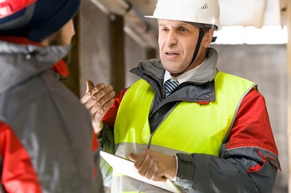 Corporate executive speaks to MRO manager on warehouse floor, both men wearing hard hats, safety vests, and winter jackets.