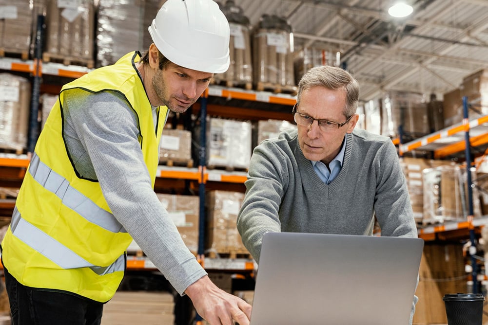 Two supply chain professionals reviewing MRO procurement and inventory data on a laptop inside a warehouse facility.